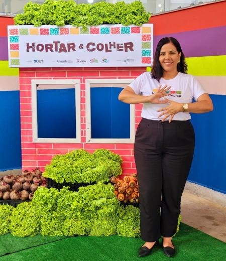 Educadora diante da horta cenográfica e dos alimentos colhidos no projeto Hortar e Colher